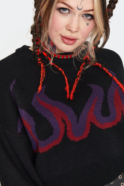 a model wearing a thick jacquard knit sweater with an oversized cropped fit. It has a purple and red flame pattern on a black background across the chest and sleeves. Shown from front in close up