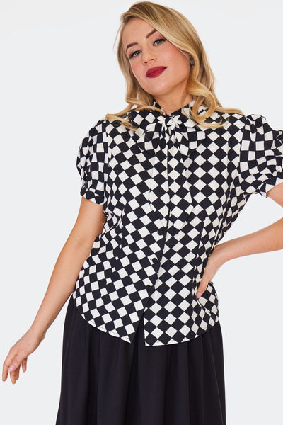 A model wearing a short sleeved tie neck blouse in a bright white and black diamond harlequin pattern. It has puffed sleeves and black fabric covered buttons. Shown from the front untucked