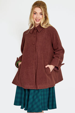 a model wearing a warm brown swing coat made of a thick textured faux wool. It has a pointed fold over collar, slash pockets, three self buttons, and sleeves with notched cuffs. Shown closed from front