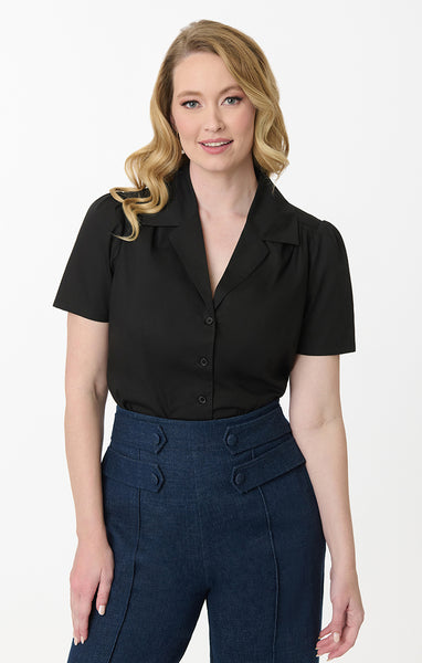 model wearing black cotton short sleeve blouse with a notched collar, slightly puffed shoulders, and matching black plastic buttons. Shown from front tucked into pants