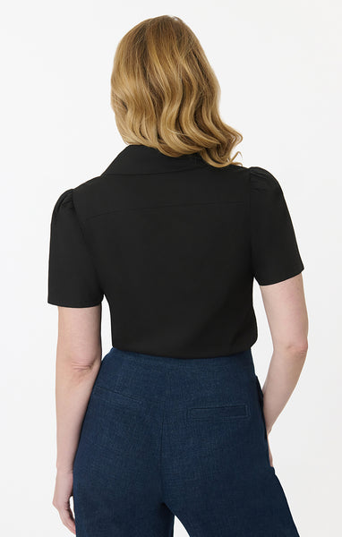 model wearing black cotton short sleeve blouse with a notched collar, slightly puffed shoulders, and matching black plastic buttons. Shown from back tucked into pants