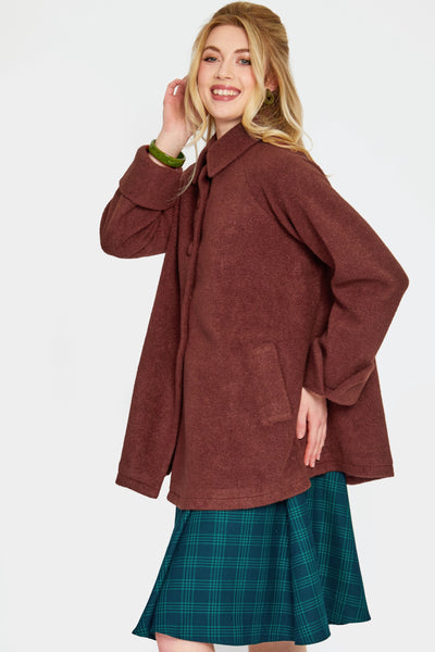 a model wearing a warm brown swing coat made of a thick textured faux wool. It has a pointed fold over collar, slash pockets, three self buttons, and sleeves with notched cuffs. Shown closed from side