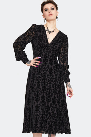 a model wearing a below the knee length dress in burnout black velvet with a damask pattern. It has a v-neckline, decorative rose-shaped black plastic buttons, and gathered long semi-sheer sleeves with button cuffs. Shown from the front