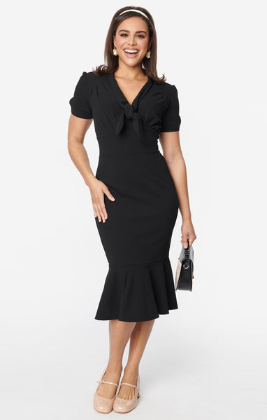 A model wearing a below the knee length black wiggle dress with a tie detail at the v-neckline, slightly puffed short sleeves with button details, a gathered underbust hem, and a mermaid hem skirt. Shown from the front