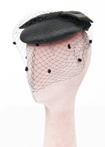 Black satin rounded pillbox style hat in satin with flat self bow on top and front netting in black with flocked dots. Shown from front on mannequin head