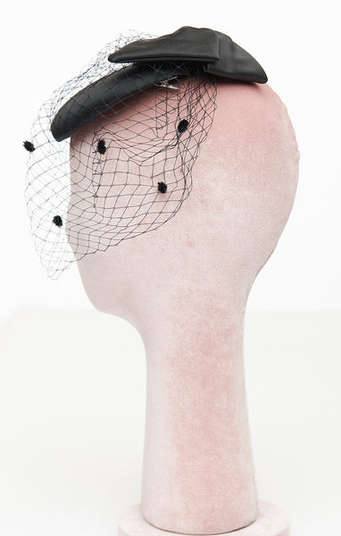 Black satin rounded pillbox style hat in satin with flat self bow on top and front netting in black with flocked dots. Shown from side on mannequin head