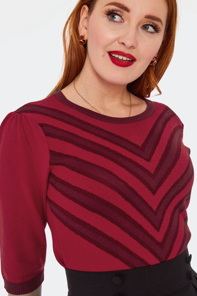 A model wearing a medium red pullover sweater with a vertical chevron pattern in a darker shade of red with matching collar and 3/4 sleeves. Seen from front tucked in close up