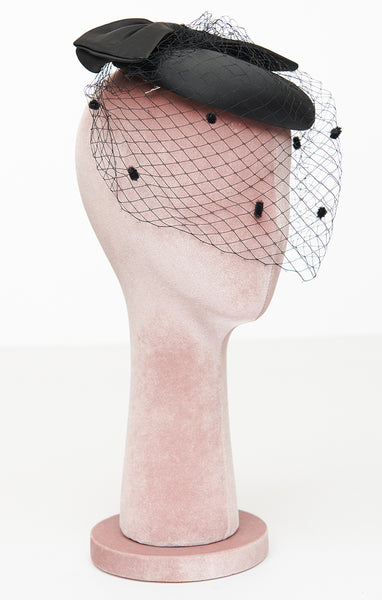 Black satin rounded pillbox style hat in satin with flat self bow on top and front netting in black with flocked dots. Shown from front on mannequin head