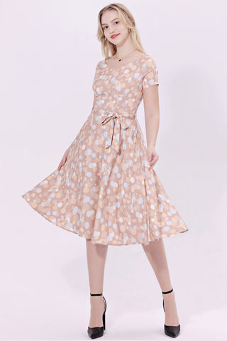 Model wearing a cocktail length short sleeved dress with a full gathered skirt and surplice style v neckline. It has a champagne beige background patterned with irregular dots in bone white, powder blue, orange, and beige. Shown from front