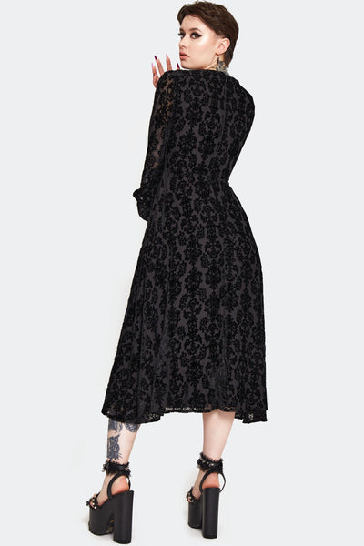 a model wearing a below the knee length dress in burnout black velvet with a damask pattern. It has a v-neckline, decorative rose-shaped black plastic buttons, and gathered long semi-sheer sleeves with button cuffs. Shown from the back