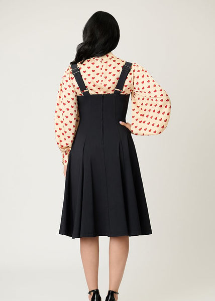 Model wearing black woven swing skirt with attached adjustable suspenders and patch pockets in the shape of red hearts with white scalloped borders. Shown from back