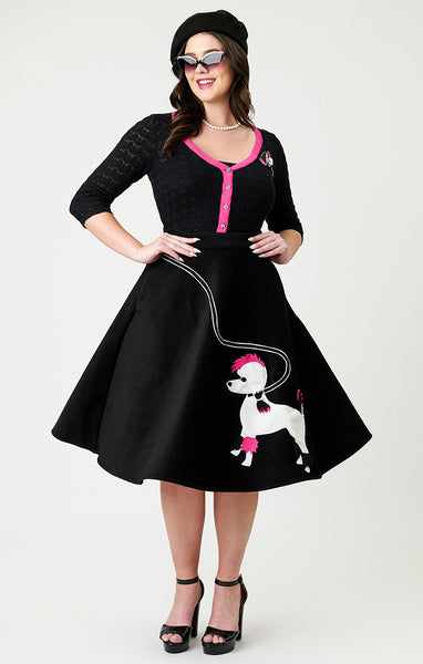 model wearing black felt style fabric swing skirt with pink, white, and black felt applique of Gonk, Elvira's poodle at bottom left hem with black and white leash applique running to right hip of skirt. Shown from front 