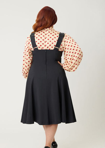 Model wearing black woven swing skirt with attached adjustable suspenders and patch pockets in the shape of red hearts with white scalloped borders. Shown from back