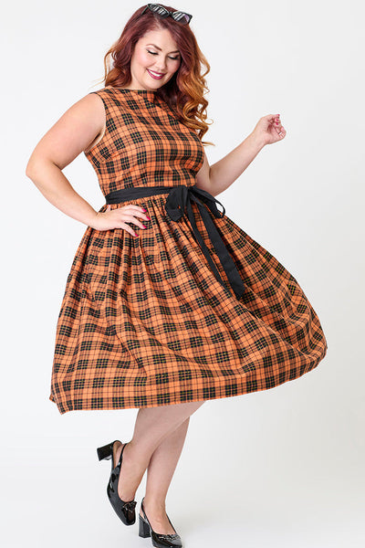 Plus size model wearing a cotton orange, black, and yellow gold lurex plaid patterned swing dress with slash neckline, darted bodice, black cotton sash belt, and below the knee gathered skirt. Shown from front with skirt twirling