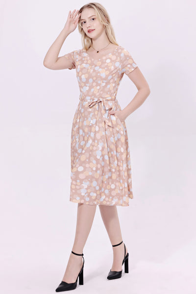 Model wearing a cocktail length short sleeved dress with a full gathered skirt and surplice style v neckline. It has a champagne beige background patterned with irregular dots in bone white, powder blue, orange, and beige. Shown from front