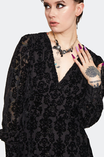 a model wearing a below the knee length dress in burnout black velvet with a damask pattern. It has a v-neckline, decorative rose-shaped black plastic buttons, and gathered long semi-sheer sleeves with button cuffs. Shown from the front in close up
