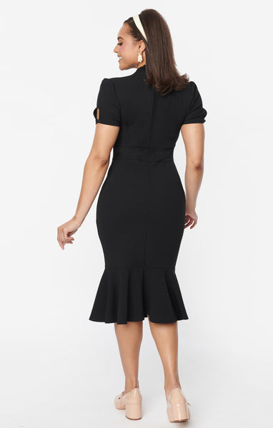 A model wearing a below the knee length black wiggle dress with a tie detail at the v-neckline, slightly puffed short sleeves with button details, a gathered underbust hem, and a mermaid hem skirt. Shown from the back