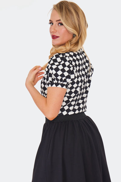 A model wearing a short sleeved tie neck blouse in a bright white and black diamond harlequin pattern. It has puffed sleeves and black fabric covered buttons. Shown from the side