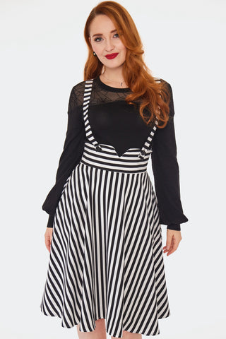 A model wearing a below the knee length swing skirt in a black and white vertically striped pattern. It has matching suspender style straps attached with two black buttons at the front of the high cummerbund style notched waistband. Shown from the front