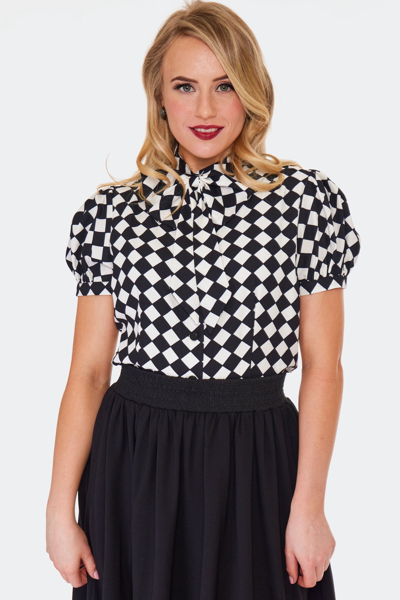 A model wearing a short sleeved tie neck blouse in a bright white and black diamond harlequin pattern. It has puffed sleeves and black fabric covered buttons. Shown from the front tucked into a skirt