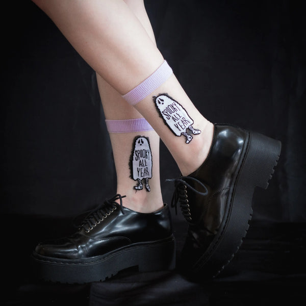 A pair of crew socks with a pale purple cuff, toe, and heel with the image of a ghost with the words “SPOOKY ALL YEAR” written on the middle of the ghost’s sheet. The brand name Ectogasm is written on the bottom of the sock , shown worn by model