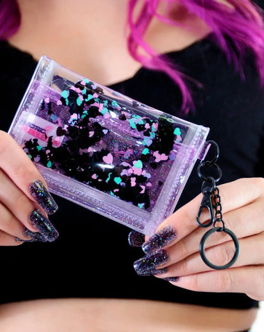 A model holding a small rectangular wallet made of clear and purple sparkly vinyl with pockets of liquid multicolored glitter. It has a gunmetal keychain ring