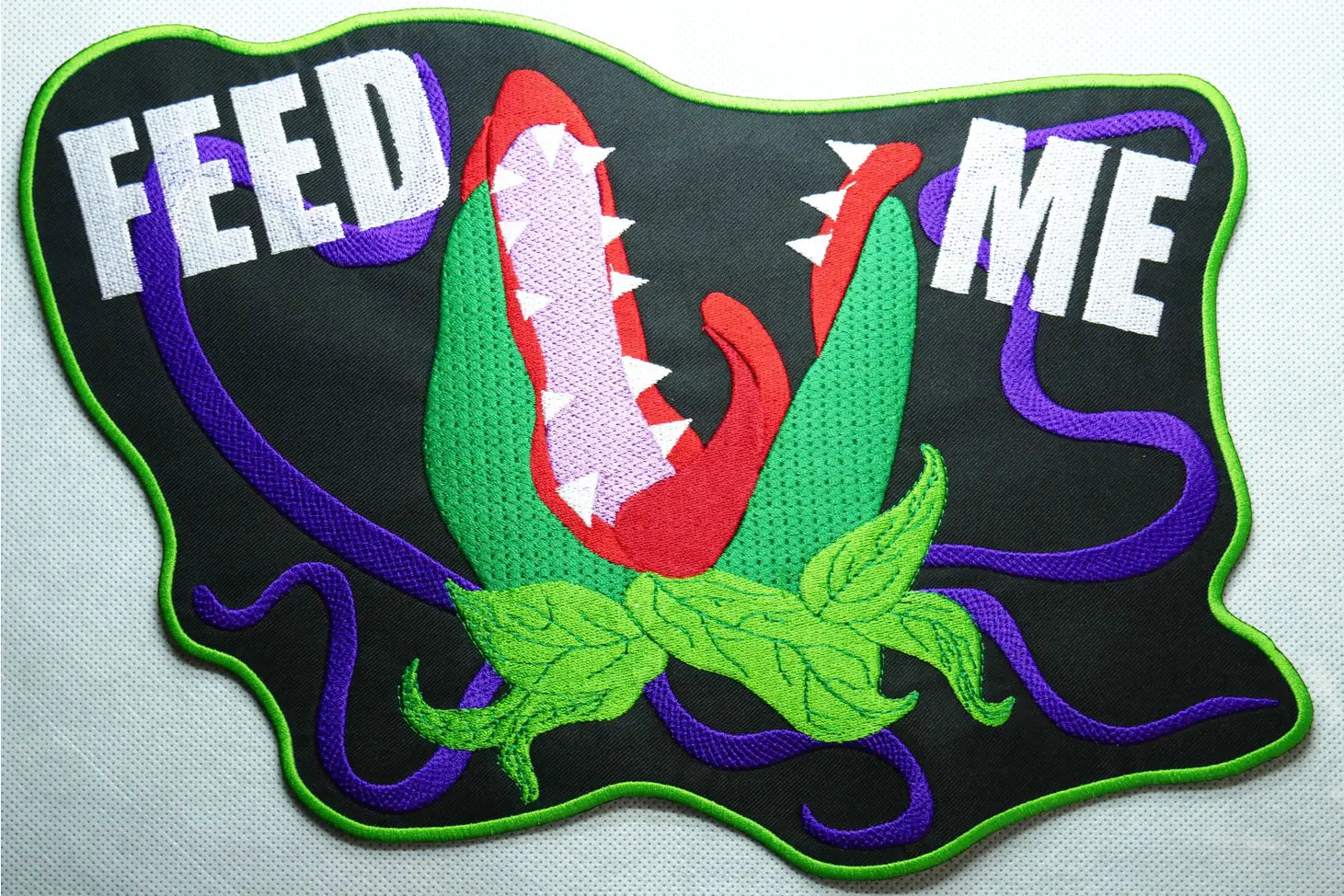 A back patch of Audrey 2 from Little Shop of Horrors in green, red, and purple with white "FEED ME" text embroidered on a green-bordered black twill patch.