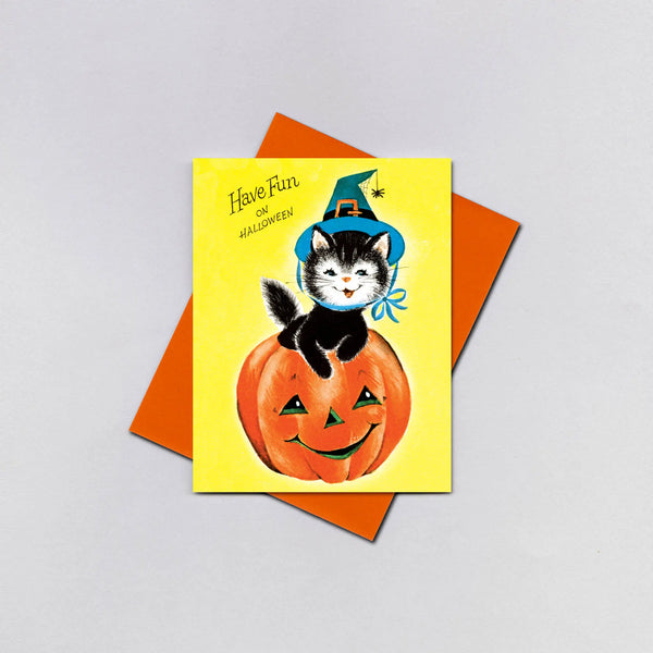 A rectangular greeting card with a black kitten sitting in a jack-o’-lantern wearing a blue witch hat in front of a yellow background. It has the message “Have fun on Halloween!”