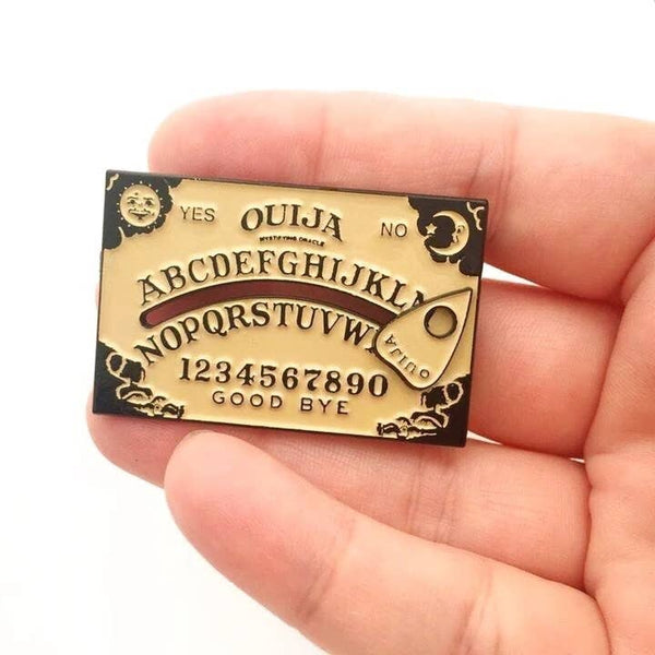 black nickel metal and tan enamel Ouija Board lapel pin with attached sliding planchette, held in a hand