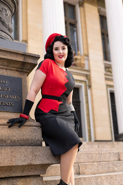 A 40s style short sleeved dress with sawtooth style color blocking in red and black down the front. It has dolman-style cap sleeves, red buttons down the length of the dress, and a matching fabric belt in black with a plastic buckle. Shown on a model