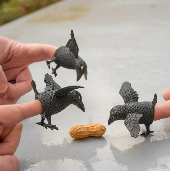 three 2" soft vinyl black crow finger toppers shown on hands and pecking at a peanut