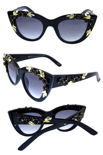 Thick black plastic cat-eye sunglasses with striking ornate gold metal leaf and black floral details on both the frame front and arms, showing three pairs in different views