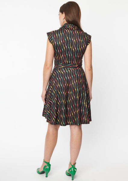 A fit and flare dress with a cowl neck and cap sleeves. It has a pattern of swirled dots in red, green, yellow, and blue on a black background. Shown on a model from behind