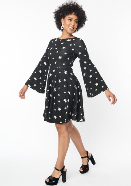 A black sweater knit fit and flare dress with raglan bell sleeves in a black and white star pattern. Shown on a model with their arms posed to display bell sleeves