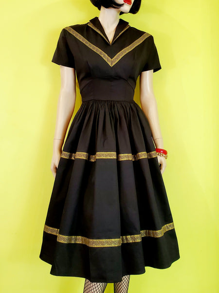50s style Patio Dress in black stretch cotton sateen embellished with metallic gold ribbon trim features a collared v-neckline, fitted princess seamed bodice, short dolman sleeves, and gathered full  just-below-the-knee length skirt. shown on mannequin