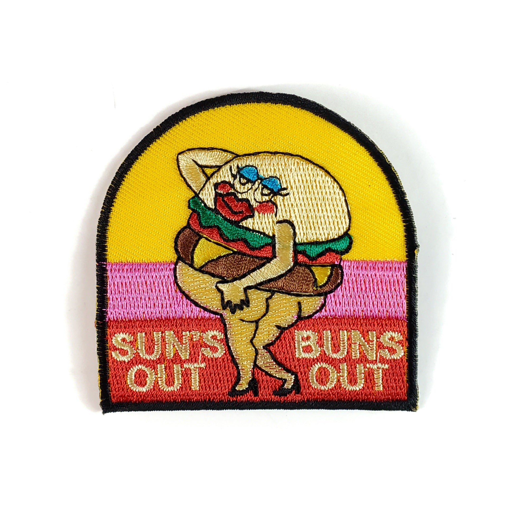 A semicircular embroidered patch of a hamburger with a human face, butt, and legs in high heels posing behind a yellow, pink and red background. The words “SUN’S OUT BUNS OUT” are written at the bottom of the patch