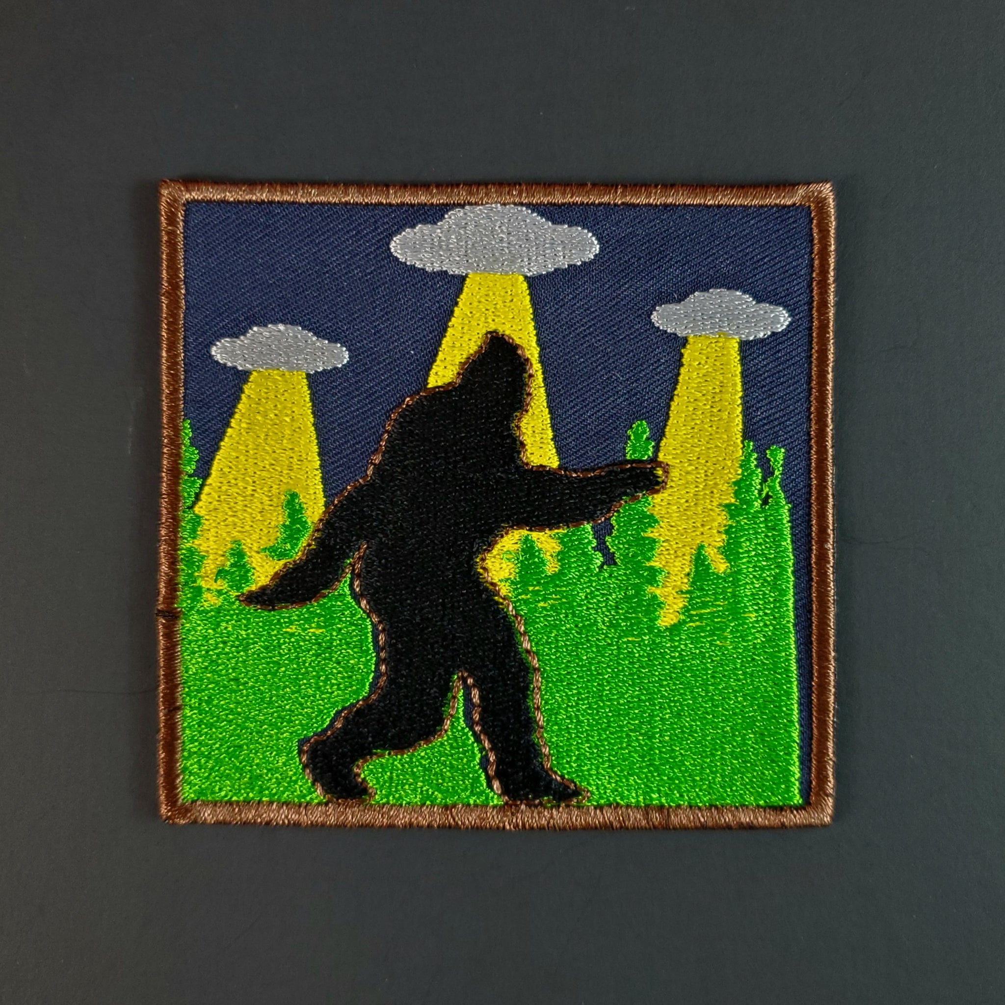 embroidered patch of Sasquatch walking silhouette against background of green forest trees and three UFOs in the night sky