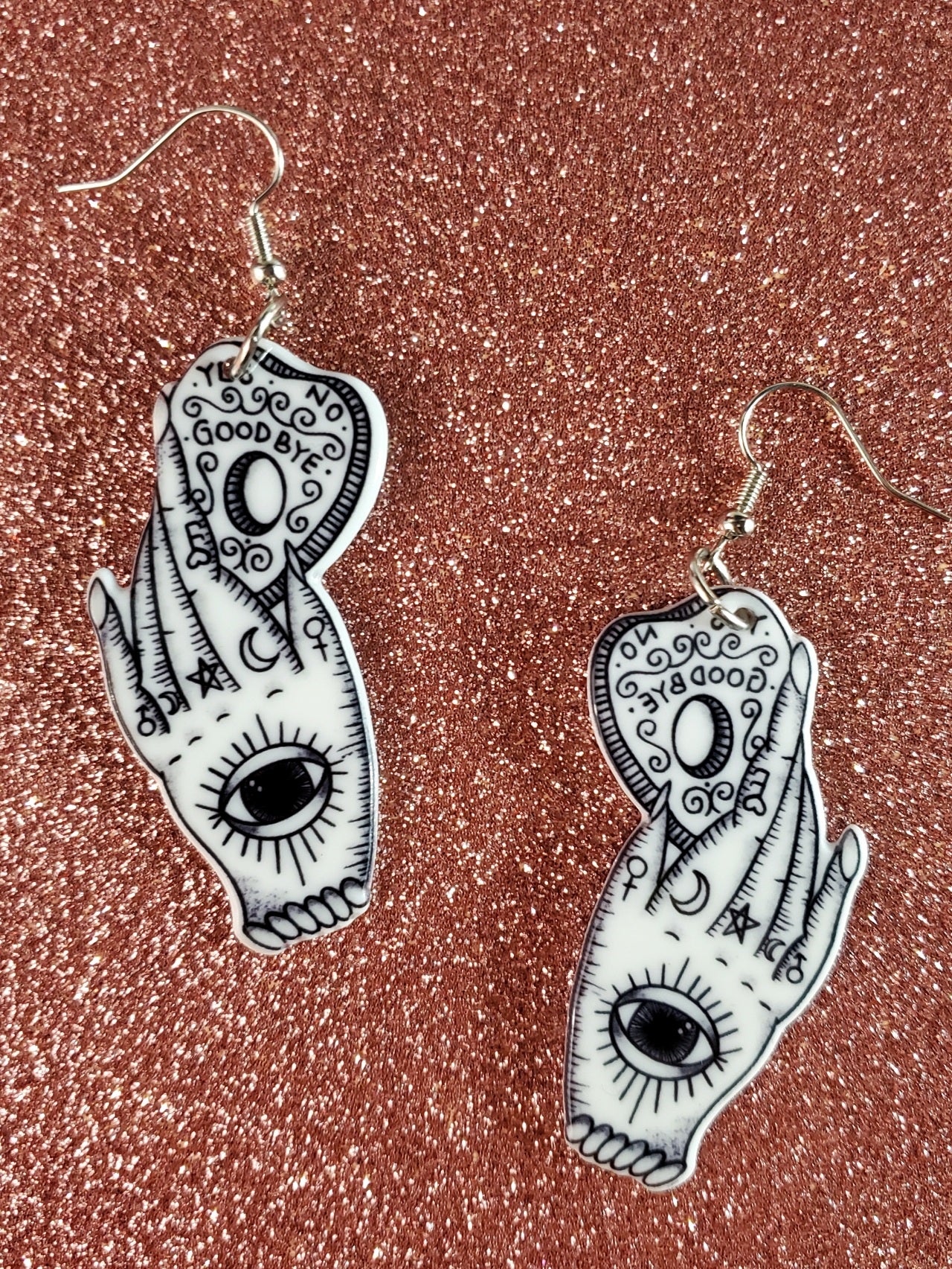 A pair of white acrylic coated earrings with an image of a hand covered in occult symbols with a large eye on its palm holding a ouija board planchette