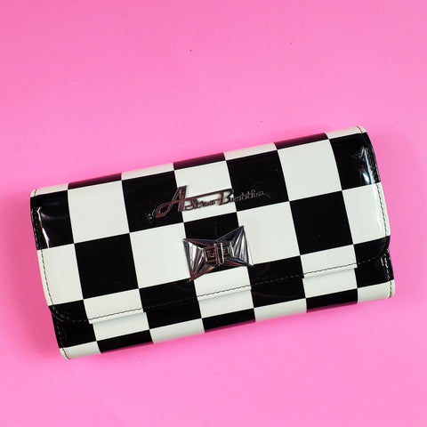 A rectangular tri-fold wallet in a shiny black and white checkerboard vinyl pattern. It has a large silver Astro Bettie logo on its exterior with a silver metal clasp in an Art Deco style 