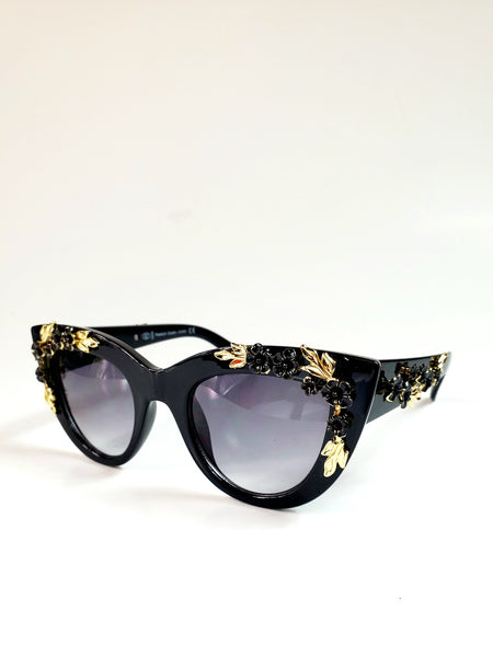 Thick black plastic cat-eye sunglasses with striking ornate gold metal leaf and black floral details on both the frame front and arms, showing 3/4 view