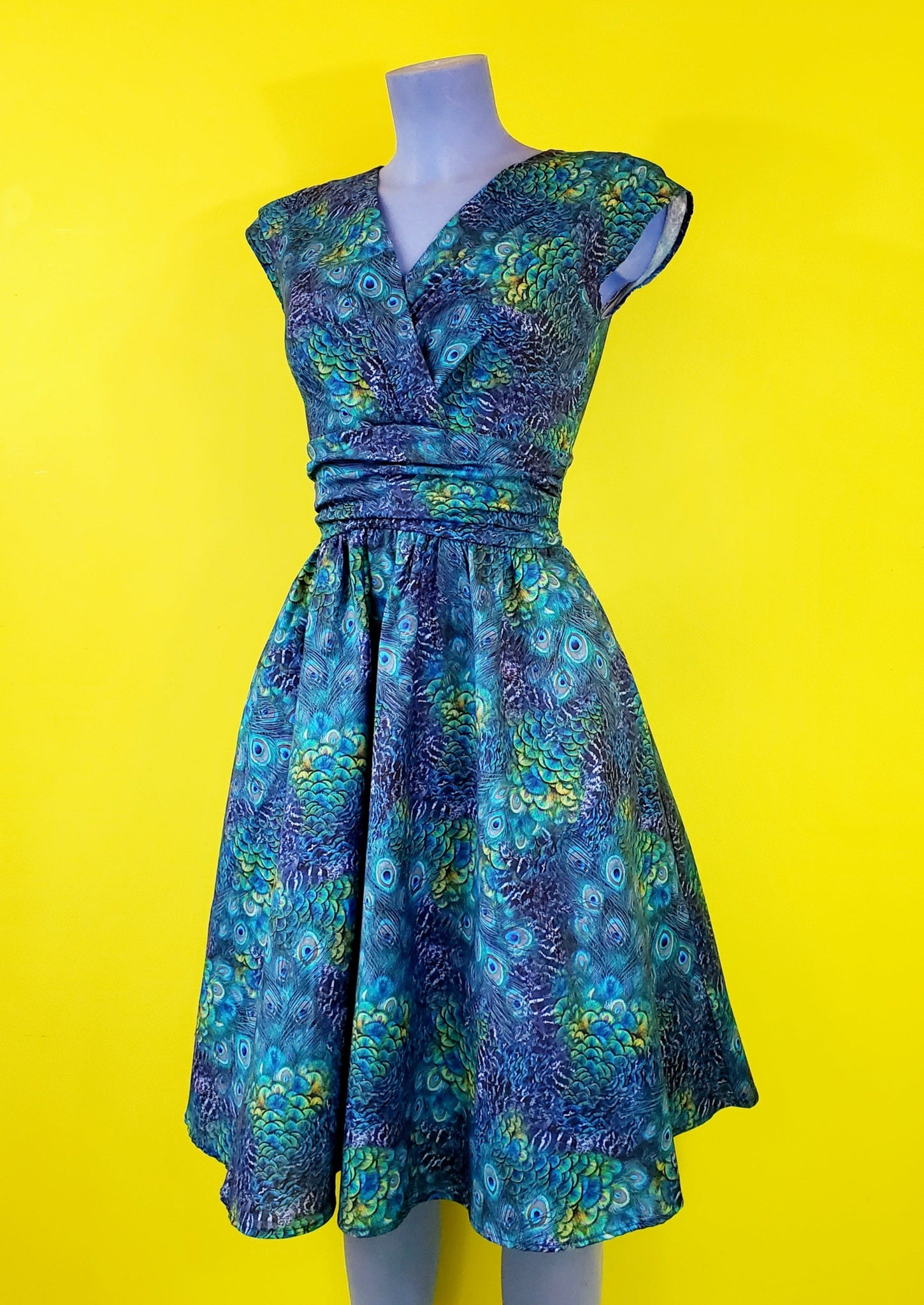 cap sleeve cotton dress in a green and blue peacock feather print with a surplus gathered neckline, wide gathered waist, and a just past the knee full skirt, shown on a mannequin