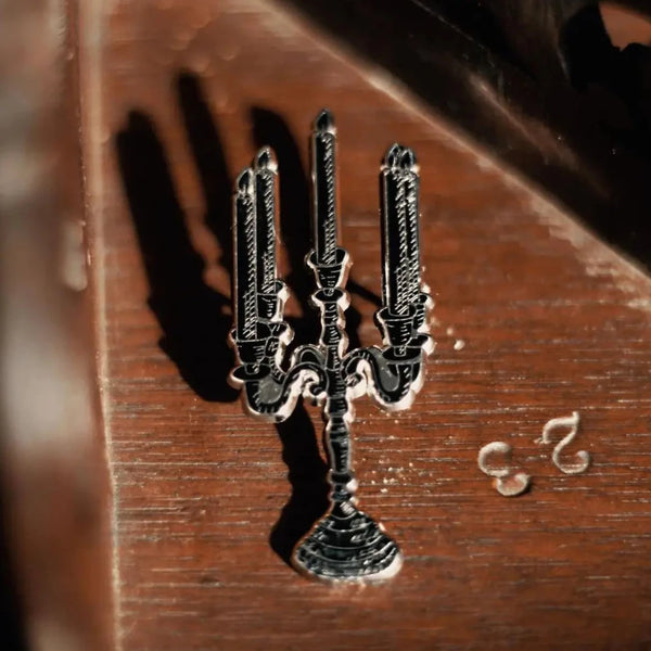 An enamel pin of a silver and black candelabra with five candles, shown in close up
