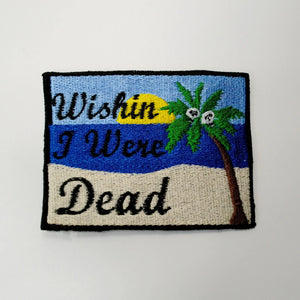 An embroidered patch in the style of an old fashioned postcard. “Wishin I Was Dead” in script lettering in front of a beach scene with setting sun and a palm tree with skulls in place of coconuts