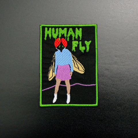 An embroidered patch depicting a woman with a fly’s head wearing a blue shirt, pink miniskirt, and white boots. On a black background with green border and green text in a dripping style font that reads “Human Fly”
