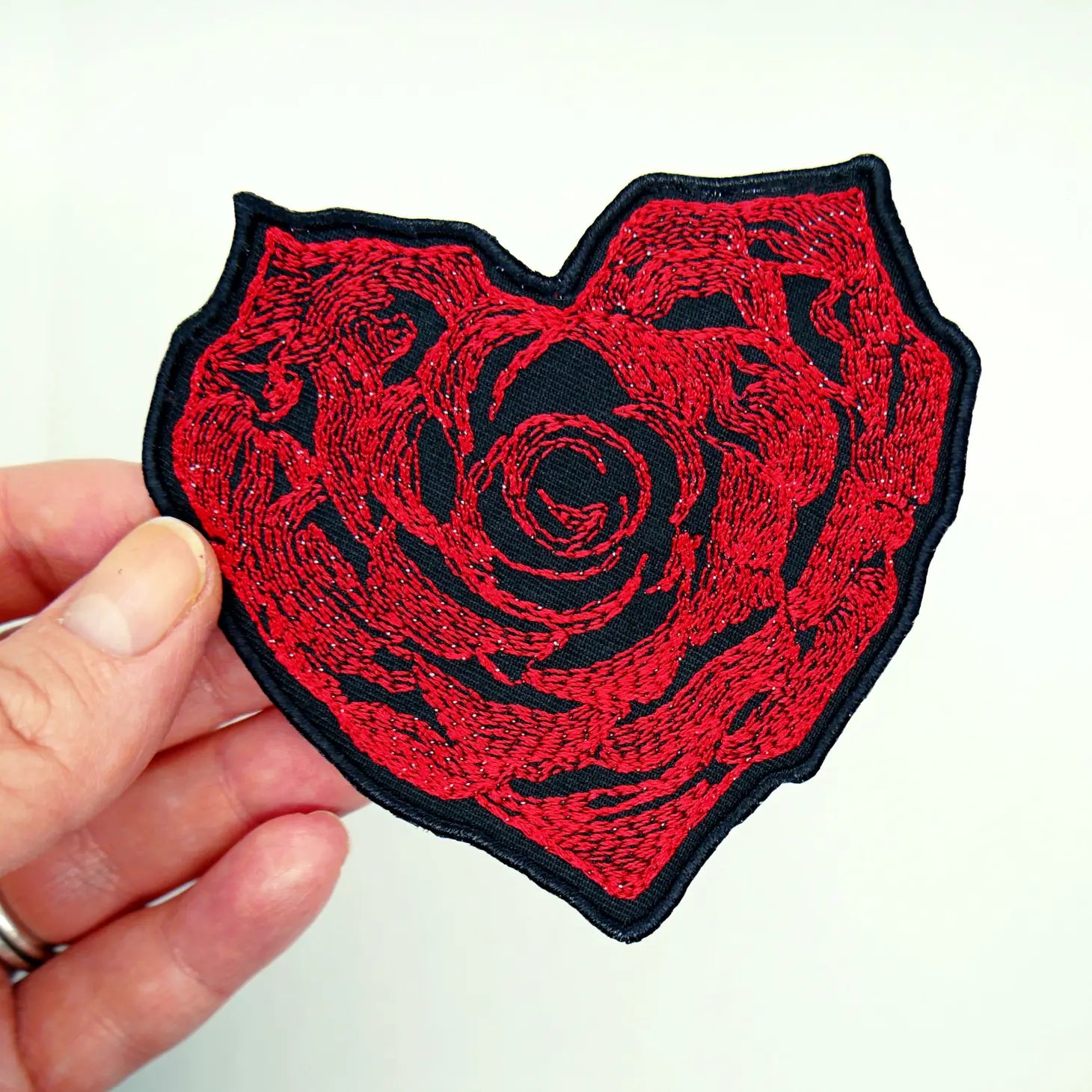 An intricate red rose embroidered patch in the shape of a heart on a black background. Held by a hand
