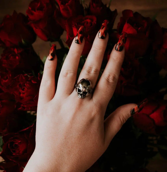 A stainless steel ring of a detailed skull on a solid stainless steel band. Shown on a hand with long black and red fingernails in front of a bouquet of red roses