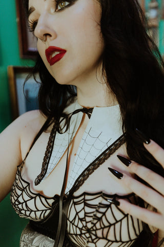 A dagger-style detachable collar that is made of soft matte white mercerized cotton and has a black satin ribbon tie. The front of the collar has delicate black spiderweb embroidery and a black lace trim. Shown on a model in close up