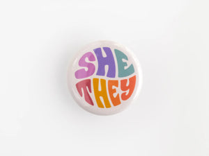 A 1.25” button with the pronouns “she” and “they” in multicolored lettering