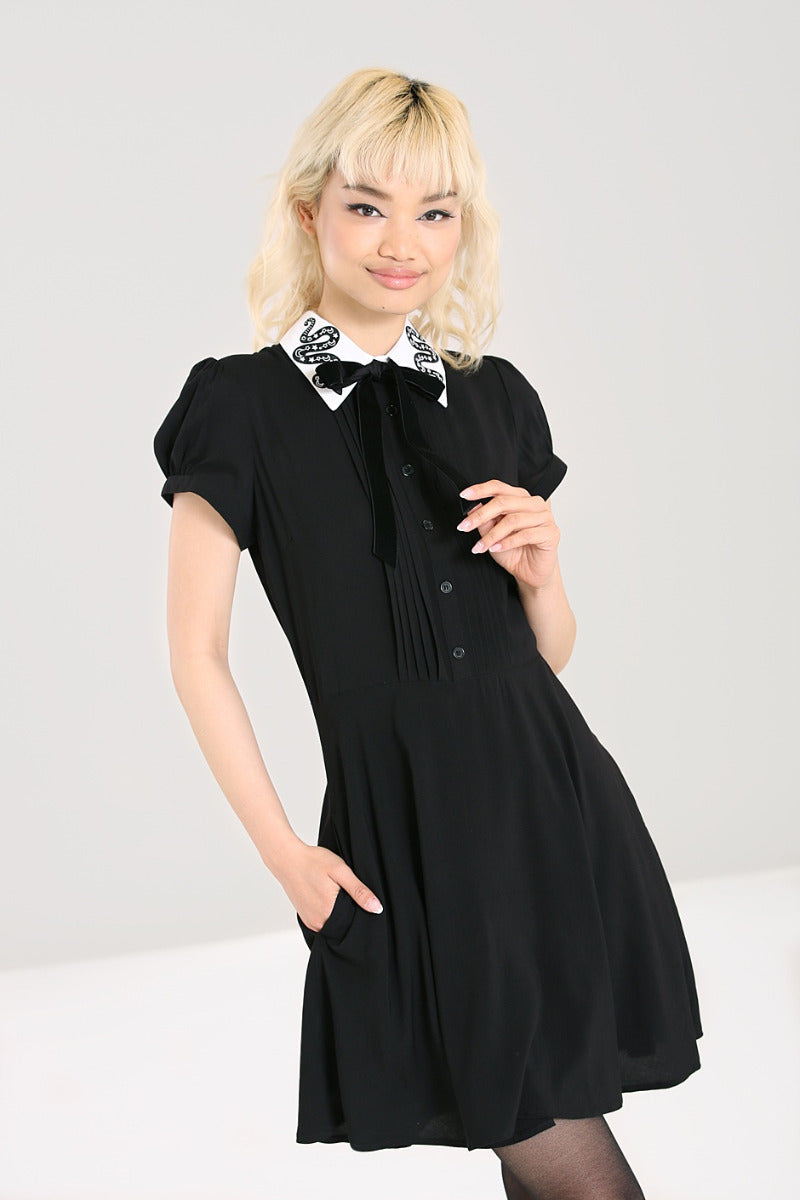 A shirtwaist mini dress with a white pointed collar that has stylized snake embroidery on each side. The dress has a black velvet bow tied around the neck and pin tuck details down the bodice in between the buttons. The sleeves are short and slightly puffed. Shown on model with one hand in pocket 