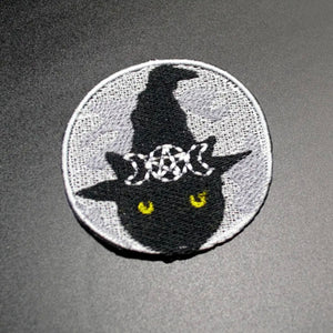 embroidered patch of a black cat wearing a witch hat surrounded by three moons and a pentagram on a grey background with matching clouds